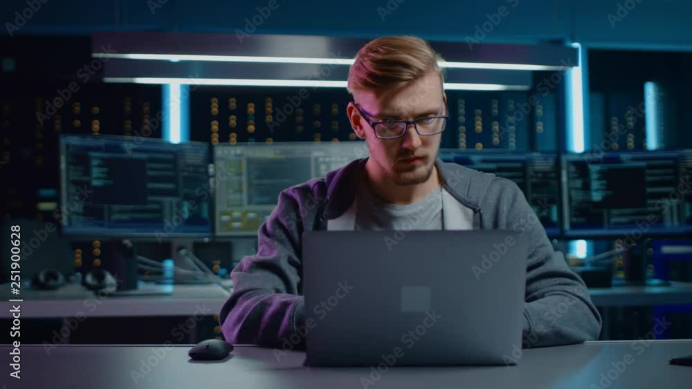 Poster Portrait of Software Developer / Hacker / Gamer Wearing Glasses and Headset Sitting at His Desk and Working / Playing on Laptop. In the Background Dark High Tech Environment with Multiple Displays.