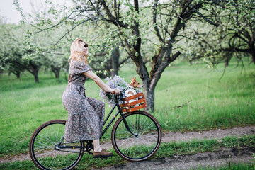 blonde girl in a dress and glasses rides a bicycle with a basket with a dog and flowers in a flowered garden
