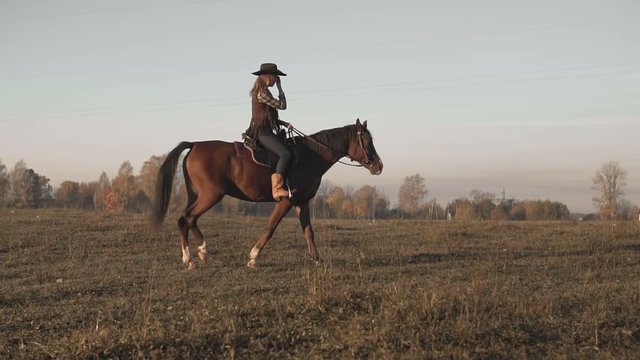 Young cowgirl at brown horse in slow motion outdoors. Beautiful woman riding horse in background sunrise in field