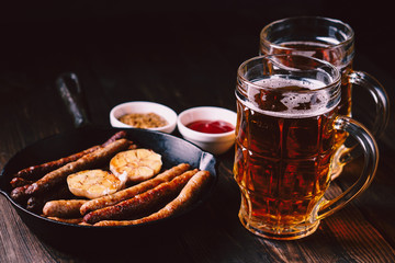 beer and appetizing snacks. table with two mugs of craft lager and frying pan with homemade grilled sausages. oktoberfest food, pub concept