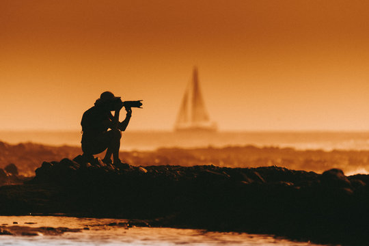 Silhouette of a photographer at the sunset sitting near the sea. Man with photo camera taking pictures at the beach. Sail boat in the background Orange sky and waves. Summer vacation evening landscape