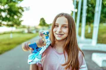 Teenager girl 10-12 happy smiling in freckles and with braces on teeth, in summer in the city, on the shoulder a skateboard for skating. Long hair, pink t-shirt.