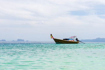 Small wooden tourist boat anchoring on the beach with view of white cloudy sky, island and green clear sea water in Krabi, Southern Thailand.