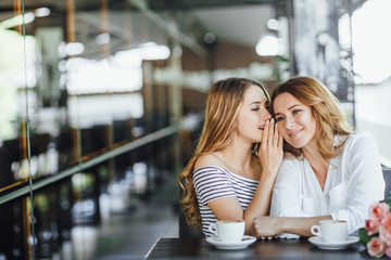 A young beautiful blonde girl speaks to her mom an interesting secret, they sit on a summer terrace cafe, happy and laugh.