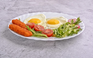 Breakfast. Fried eggs with vegetables and fish fingers. Tomato, green salad, radish, fish fingers, fresh dill and two eggs on a white plate.