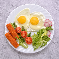 Breakfast. Fried eggs with vegetables and fish fingers. Tomato, green salad, radish, fish fingers, fresh dill and two eggs on a white plate. Top view.