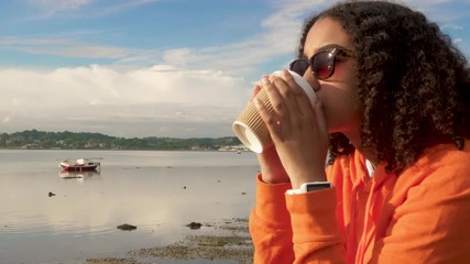 Beautiful mixed race African American girl teenager young woman wearing sunglasses and an orange hoodie, drinking takeout coffee by a harbor looking sad or thoughtful