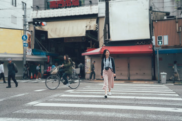 vintage style photo of full length young asian female photographer walking on zebra cross street crosswalk. people riding bike in background. Translation on wall building text "kuromon market".