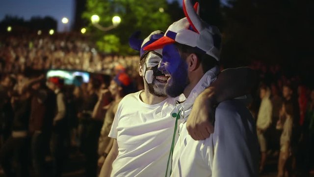 Soccer fan are gay with paint face hugging playfully watching match. People from LGBT community stand stadium amid fan, are smiling each other cutely. Love couple guy embracing watching football match