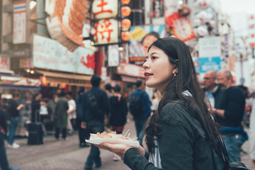 vintage photo of young girl tourist standing on dotonbori street outdoor holding local specialty snack. asian woman sightseeing in teeming shopping area in famous tourism attraction trying takoyaki. - Powered by Adobe