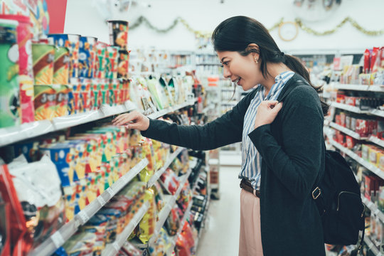 sale shopping consumerism people concept. asian woman backpacker with curious face picking snack cookies choose at grocery supermarket. lady buying products food instant noodles in convenience store.
