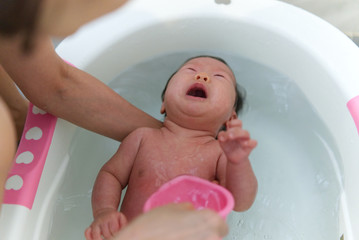 Asian mother take a bath her newborn baby in bath tub and rinsing the soap from the body in bathroom. Mother holding the child with care but the child is a little bit crying.