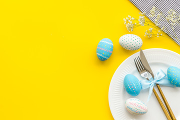 Easter table decoration. Plate, cutlery, painted eggs and dry white flowers, tablecloth on yellow background top view copy space