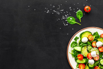 Fresh salad with mozzarella, spinach, cherry tomatoes, cucumber on plate on black background top view space for text