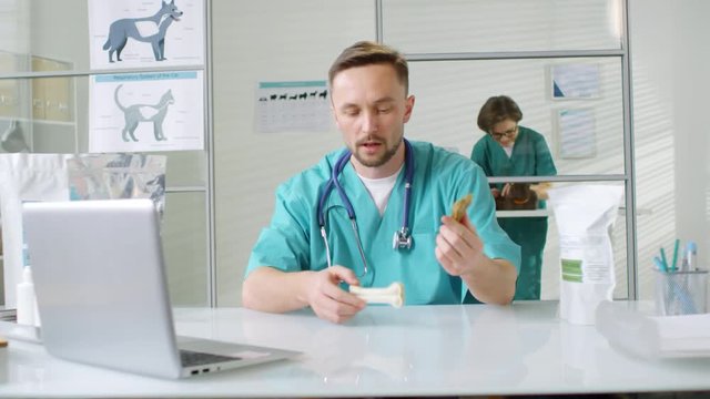 Medium shot of male vet sitting at laptop and giving online comments about dog chew bones to customer, while his female assistant examining cat on background