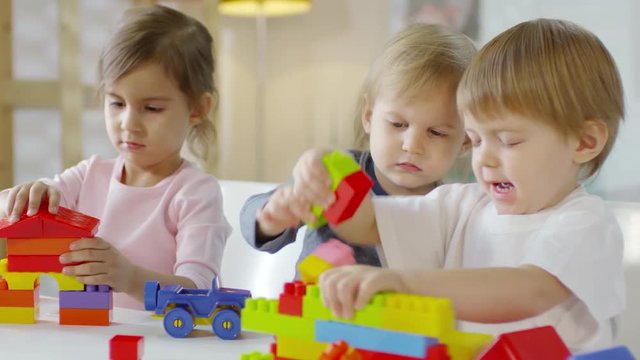 Two little caucasian boys and pretty girl playing with educational construction toys at table in kindergarten