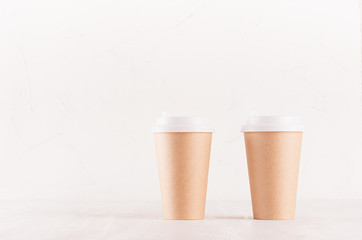 Coffee packing mockup -  two kraft brown paper cups with white cap on white wood board with copy space, coffee shop interior. Modern elegant concept for branding identity, advertising, design.