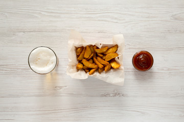 Fried potato wedges in paper box, barbecue sauce and glass of beer on a white wooden background. Flat lay. Overhead, top view, from above.
