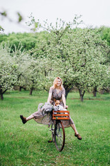 girl blonde in a dress rides a bicycle with a basket with a dog in a blooming garden