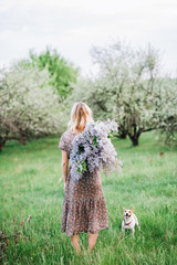 blonde girl in a dress stands with her back with a bouquet of lilac near the dog on the background of a green garden