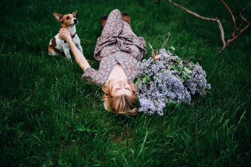 blonde girl in a dress lies on the green grass near the bouquet with lilac and hugs the dog