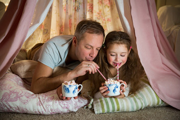 Family quality time. Father and daughter lie in homemade pink tent with flowers, drinking hot...