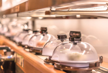 Close-up on Japanese Conveyor belt sushi restaurant where plates with sushi are placed on a rotating rail protected with a clear plastic lid. - Powered by Adobe