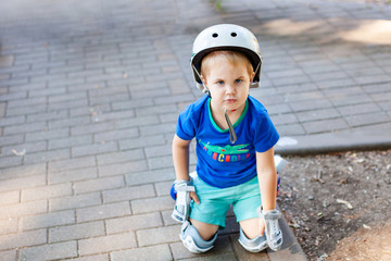 Little blonde boy 3 years old in white sport helmet and blue t-shirt riding on the roller-slates in the summer green park. Special problems with kid's eyes. Myopie, astigmatism, cross-eyed