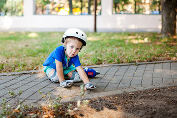 Little blonde boy 3 years old in white sport helmet and blue t-shirt riding on the roller-slates in the summer green park. Special problems with kid's eyes. Myopie, astigmatism, cross-eyed