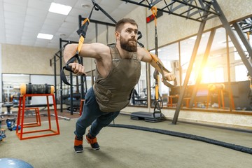 Muscular bearded man dressed in military weighted armored vest doing exercises using straps systems...