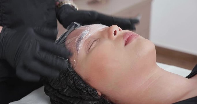 Applying plastic foil on numbing cream for eyebrow tattoo removal. Female cosmetologist preparing to remove the permanent tattoo from eyebrows. Woman pacient in a beauty salon.