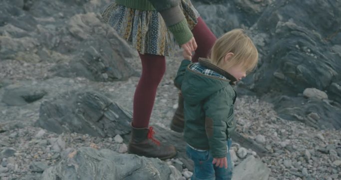 Young mother and toddler walking hand in hand on rocky beach in winter