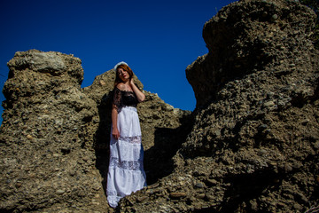 Naklejka premium Portrait of young beautiful redhead woman in dress sitting on the rock and blue sky
