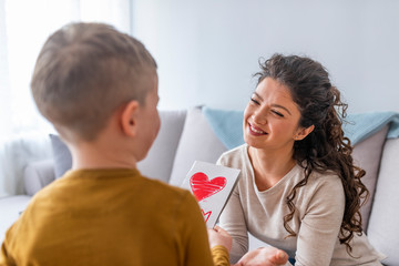 Happy mother's day! Child boy congratulates mom and gives her flowers and postcard. Mum and boy smiling and hugging. Family holiday and togetherness.