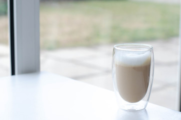 A cup of latte with froth on a table near a large window.