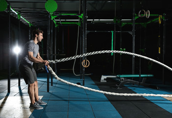 Young sporty man doing battle rope exercise in gym
