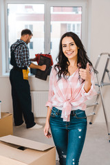 selective focus of cheerful woman showing thumb up with handyman on background