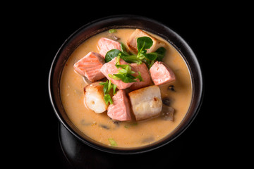 Spicy Thai soup Tom Yam with Coconut milk, Chili pepper and Seafood shrimp and salmon in a plate on a black background. Asian cuisine, restaurant menu.
