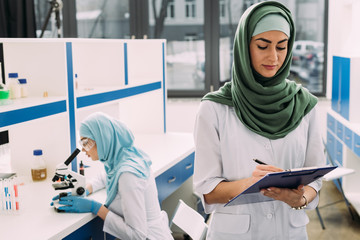 concentrated female muslim scientists using microscope and writing in clipboard during experiment...