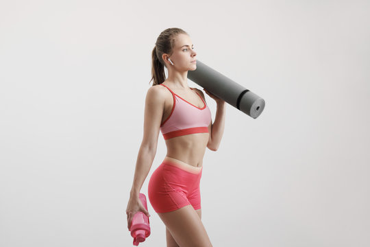 Young athletic woman with an athletic body, wearing wireless headphones and a red sportswear holding a water bottle and a fitness mat. © spaskov
