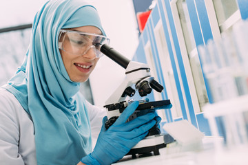 smiling female muslim scientist looking through microscope during experiment in chemical laboratory