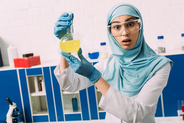 shocked female muslim scientist looking at camera and holding flask during experiment in chemical laboratory