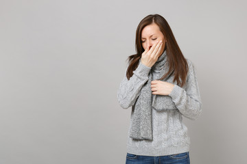 Young woman in gray sweater scarf sneezing or coughing covering mouth with palm isolated on grey wall background. Healthy lifestyle ill sick disease treatment, cold season concept. Mock up copy space.