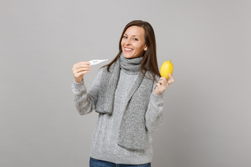 Smiling young woman in gray sweater, scarf holding lemon thermometer isolated on grey wall background in studio. Healthy lifestyle, ill sick disease treatment, cold season concept. Mock up copy space.