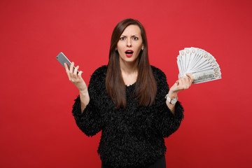 Perplexed young woman in black fur sweater holding mobile phone, fan of money in dollar banknotes, cash money isolated on red background. People sincere emotions lifestyle concept. Mock up copy space.