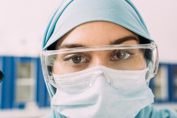 close up of female muslim scientist in medical mask, goggles and hijab looking at camera