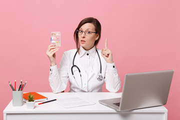 Female doctor sit at desk work on computer with medical document hold pills in hospital isolated on pastel pink wall background. Woman in medical gown glasses stethoscope. Healthcare medicine concept.