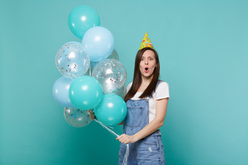 Fototapeta premium Shocked young woman in denim clothes, birthday hat keeping mouth open celebrating, holding colorful air balloons isolated on blue turquoise background. Birthday holiday party, people emotions concept.