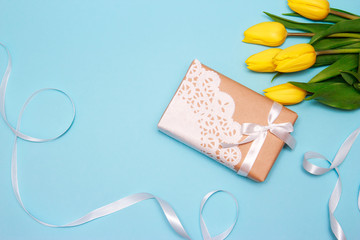 A bouquet of yellow tulips and a gift of craft paper decorated with a lace napkin on a blue paper background.. Spring background, Mothers day or Valentines day greeting card, copy space, flat lay