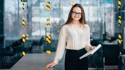 Friendly employee. Young smiling intern in glasses holding clipboard with documents stand at workplace.